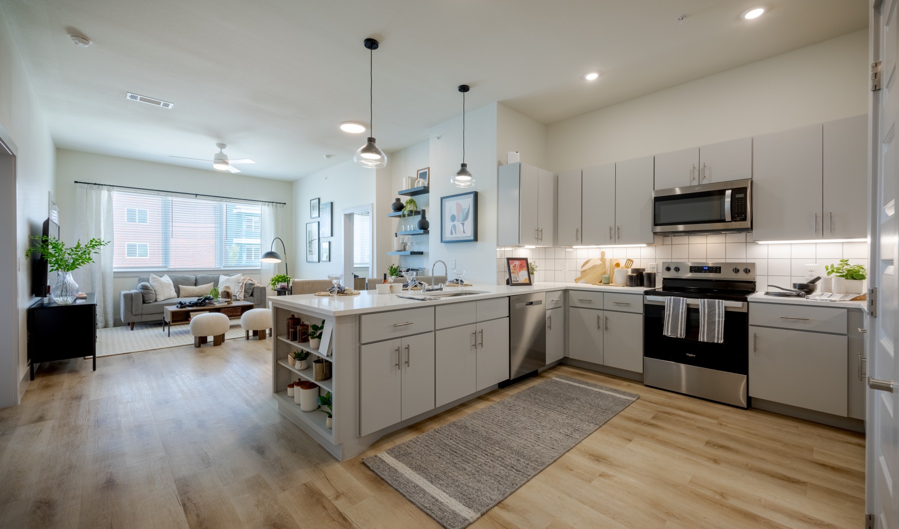 Bright, open-concept apartment kitchen and living area featuring white shaker-style cabinets, quartz countertops, stainless steel appliances, a large kitchen island with built-in shelving, and light wood flooring. Pendant lights hang above the island, whi